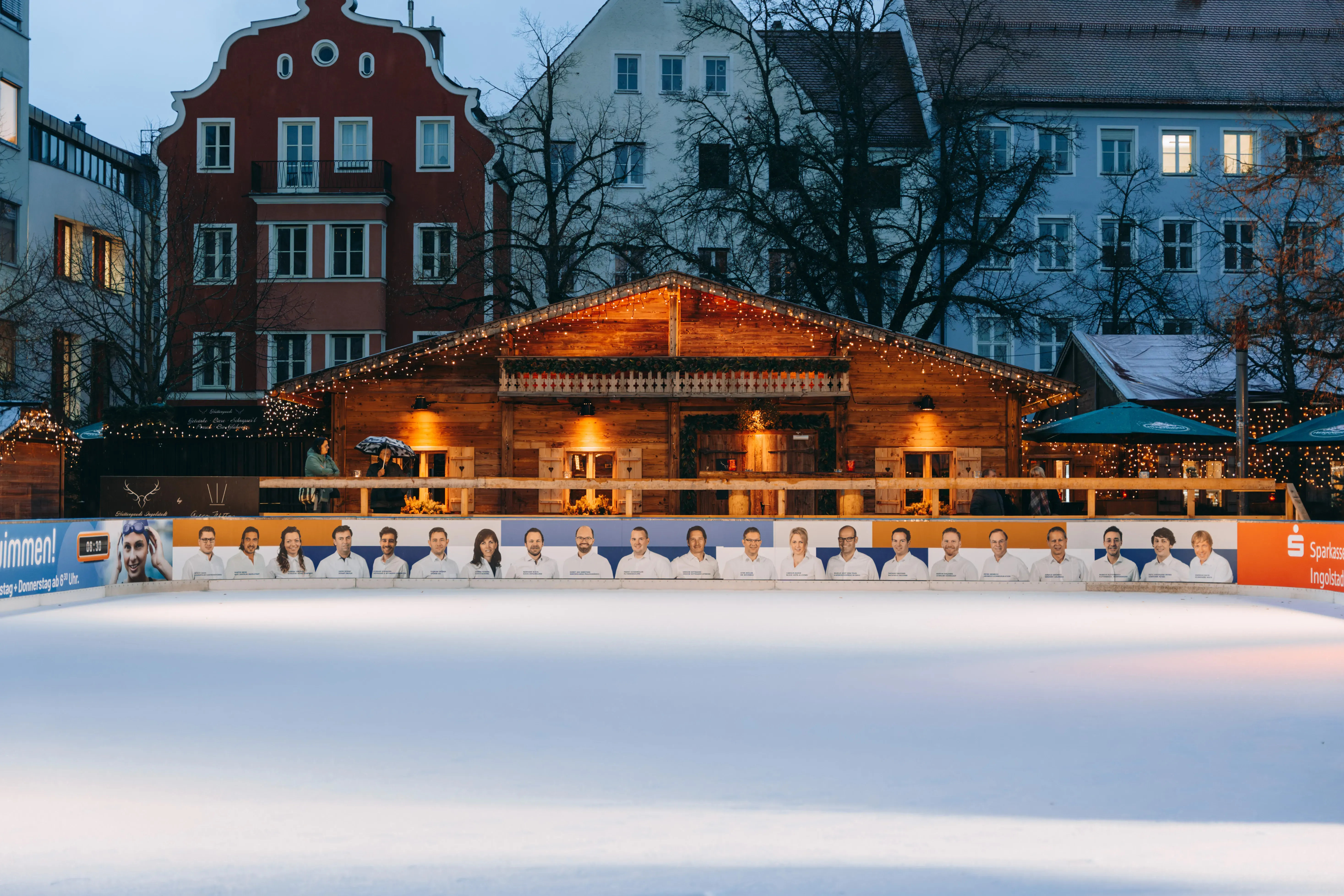 A group of people in white shirts stand on an ice rink in front of a wooden chalet decorated with lights.